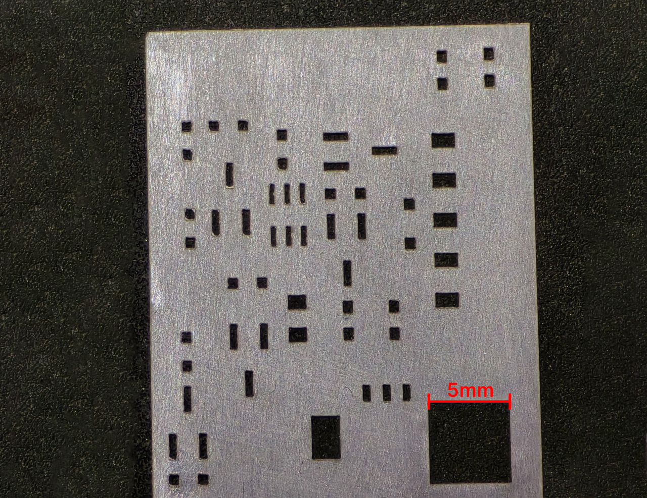 A close-up of a cut Mylar stencil with rows of small rectangular apertures and a red 5 mm scale bar for reference.