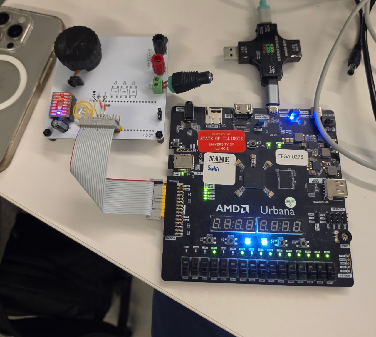 A wider view of the same bench, with the knob on its white circuit board, the gray ribbon cable, and the AMD Urbana FPGA board taking USB power through an inline USB meter.